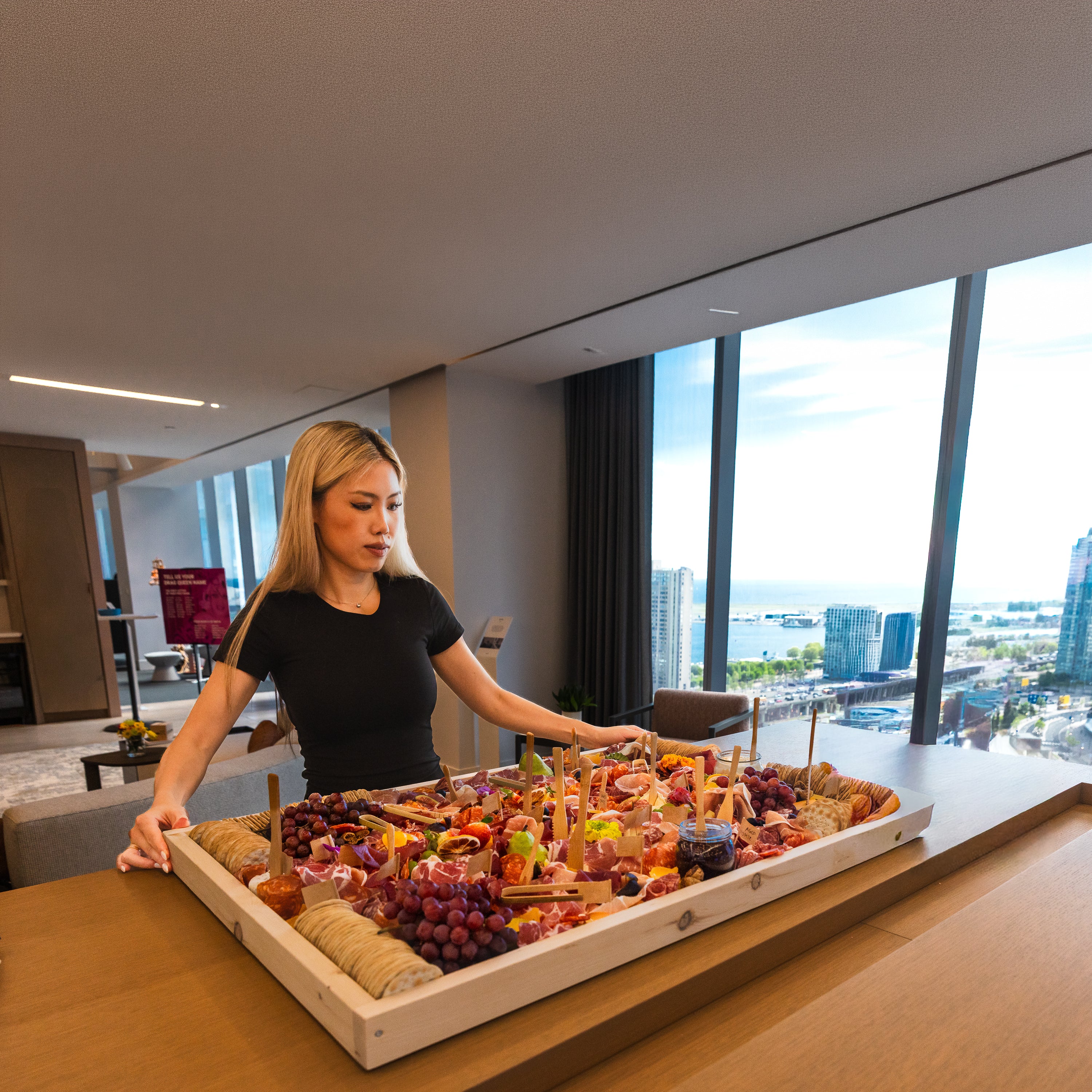 A woman in a black t-shirt is serving food from a large, elaborate charcuterie board arranged in a white tray on a wooden table in Toronto. The board is filled with various cheeses, crackers, grapes, and meats.